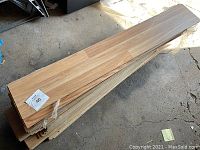 Stack of unused laminate flooring boards, light wood finish, laid horizontally under sunlight, showing natural wood grain pattern and uniform size.