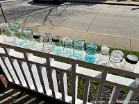 Photo of twelve antique fruit sealer jars arranged in a line on a porch railing, showing a variety of sizes, colors, and lid types.