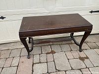 Dark wooden rectangular piano bench with a closed lid, showing carved legs and metal support braces under the seat.