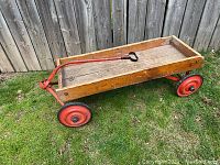 Front side view of the antique wooden wagon showing red metal frame, handle, and rubber steel wheels.