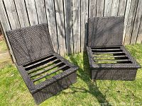 Two brown resin wicker patio loungers placed on grass in front of wooden fence, showing angled view of seats and backs with no cushions.