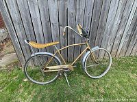 Side view of the complete gold frame banana seat bicycle leaning against a wood fence on grass