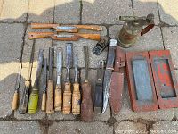 Photo showing entire lot of woodworking tools including chisels, gouges, knife with sheath, sharpening stone in wood holders, small hand plane, metal oil can, and file.