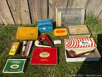 Overview of all tobacco tins, cigarette boxes, and pipes arranged on the grass against a wooden fence.