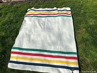 Full view of the white polar fleece blanket with Hudson's Bay Company colored stripes near the edges, laid flat on grass.