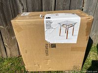 Closed shipping box containing Umbra Konnect Side Table, held outdoors on grass near wooden fence.