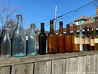 Row of sixteen antique glass bottles showing variety in color and shapes, some with wire-clasp tops and embossed lettering.
