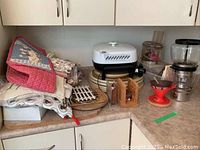 Overview of kitchen items including quilted tea cosies, oven mitts, wooden lazy susans, trivets, and electric fry pan on kitchen countertop beneath cabinets.