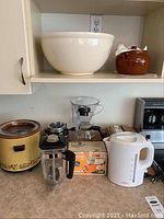 Photo shows all kitchen items together on counter: Rival slow cooker, Osterizer blender, GE mixer, Sunbeam kettle, ceramic bean pot, and large white ceramic bowl on shelf above.