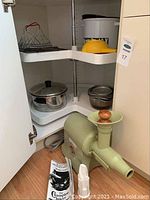 Open cabinet showing assorted pots including stock pot, double boilers, and colander along with Champion juicer on floor.