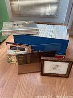 Collection of Time Life cook books stacked next to clear Plexiglas recipe stand and framed artwork with brown patterned frame.