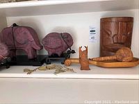Shelf with purple ceramic elephant and pig figurine, small carved wood tiki, long carved wood stick, wooden tiki drums.