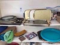 Photo showing multiple serve trays stacked on shelf, including oblong stainless steel trays with handles, round plastic trays, small decorative square trays, and woven place mats.