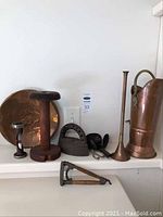View of all items in the lot including copper plate, wooden spool, flat iron, brass horn, copper pitcher, candle holder and metal tool on shelf