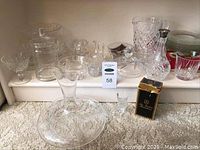 Wide-angle view showing a large clear decanter, crystal decanter with stopper, pitcher, various dessert bowls, and glasses on white shelf against beige wall.