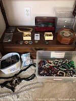 Overview photo of the costume jewelry lot showing necklaces, earrings, jewelry boxes, belts, and reading glasses on a wooden piece of furniture and on floor.
