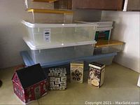 Large and medium Rubbermaid storage bins stacked, with plastic shoe boxes and several collector tins arranged in front on the floor.