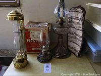 Three vintage oil lamps on a surface including a brass lamp and two glass lamps with etched chimneys. One chimney protective cover seen on side.