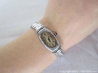 Wrist watch displayed on wrist showing oval dial with Arabic numerals and silver-tone metal expandable band.