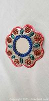 Small metal flower decoration mirror with red scalloped edges, blue central ring, orange flowers, and green leaf motifs photographed on a light surface.