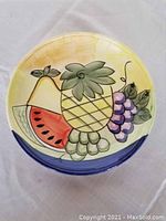 Top view of round ceramic plate with hand-painted tropical fruits: pineapple, watermelon, grapes, pear, yellow background with blue border.