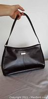 Dark brown pleather purse held by shoulder strap showing front with small silver metal plaque.