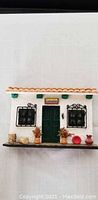 Front view of miniature white clay house with black wrought iron window bars, green door, tiled roof, and small decorative clay pots on the ledge.