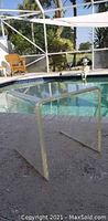 Side view of clear acrylic side table showing thickness and shape with outdoor pool background.