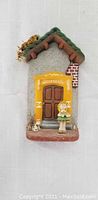 Front view of clay house figurine with detailed facade, door, girl with flowers and small dog.