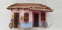 Front view of the Venezuelan purple wooden house model showing the doors, windows with wooden bars, decorative broom, wheels, and some damage to the roof tiles.
