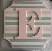Front view of wooden decorative letter 'E' sign with pastel striped background showing detail and wear.