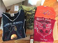 Four Hard Rock Café collectible women's T-shirts in various locations: black guitar globe design, camouflage Bangkok, orange Mexico with hummingbird, and red Las Vegas winged heart skull design.