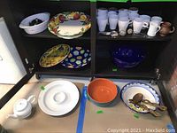 Shelf with various serving dishes, bowls, mugs, and utensils including large painted platter, bowls, and mugs.