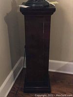 Photo showing square solid cherrywood pedestal stand placed in living room corner with a dark stain finish and minor surface lines or imperfections.