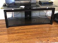 Front view of media stand showing glass top and glass bottom shelf with black frame and some electronic equipment on top (not included).