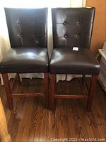 Front view of two brown leather bar stool chairs with button tufting and cherrywood legs on hardwood floor.