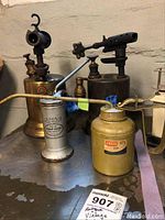 Photo of 2 antique gasoline torches (brass, with gas label and perforated nozzle), 2 vintage oil cans (yellow Marx Dunlop and silver Eagle), displayed on metal surface in workshop basement.