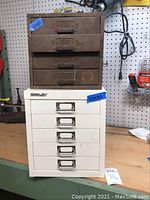 Both nuts and bolts cabinets, older rusted 4-drawer on top and newer white Bisley 5-drawer below on wood surface with pegboard background.
