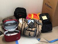Multiple bags arranged against a white wall: black backpack, brown satchel, colorful duffel bag, red and grey soft-sided bag, and black travel bags.
