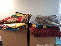 Two stacks of various ethnic blankets and tapestries on cardboard boxes including a yellow blanket, Mexican style stripy blankets, and other folded textiles.