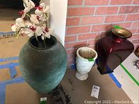 Overall view of three vases including large green clay vase with flowers, white marble-like vase, and red glass vase with gold rim.