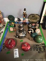 Top-down photo showing red marble apples, several ceramic spheres and eggs with various patterns and colors, a metal tea/coffee pot with wooden handle, a colorful clay island girl figurine, an Asian dragon figurine, and a brass trinket box in the background.