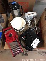 Top view photo showing all items including mandolin slicer, black and silver waffle iron, white food scale, Black & Decker hand mixer, cheese grater and Oggie thermos pitcher in cardboard box.