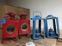 Four miniature painted oil lanterns: two red with square bodies and round windows, two blue with open-frame design and visible wick mechanism