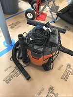 Front and side view of orange and black Rigid shop vac with attached hose and accessories lying on garage floor protected by cardboard sheets.