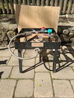 Front view of Bayou classic outdoor propane burner showing metal frame, single burner, and attached gas hose with valves. Background has wooden cutting board leaning on fence