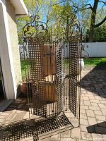 Full view of the metal 3-panel garden divider/trellis showing lattice panels and decorative top scrollwork and motifs, standing on a paved patio outside a garage.