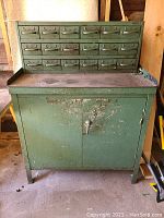 Front view of vintage green metal tool cabinet with 21 small drawers on top and two-door storage below. Paint peeling and worn from use visible.