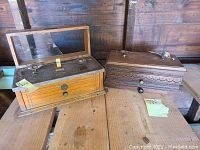 Two wooden antique scale boxes placed on wooden surface, one with glass lid and two metal scale pans visible inside, the other darker wooden box with carved decoration and drawer.
