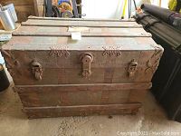 Front view of large antique trunk showing metal hardware and wood reinforced slats across the top and front face, with visible wear and missing handles.